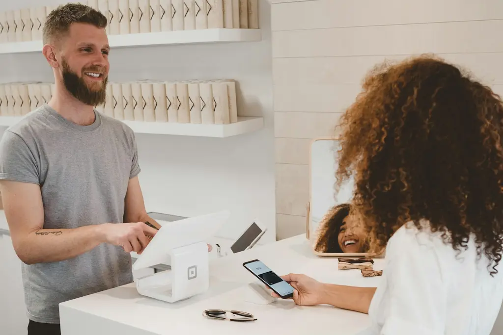 A customer paying with a phone at a shop counter while the assistant smiles