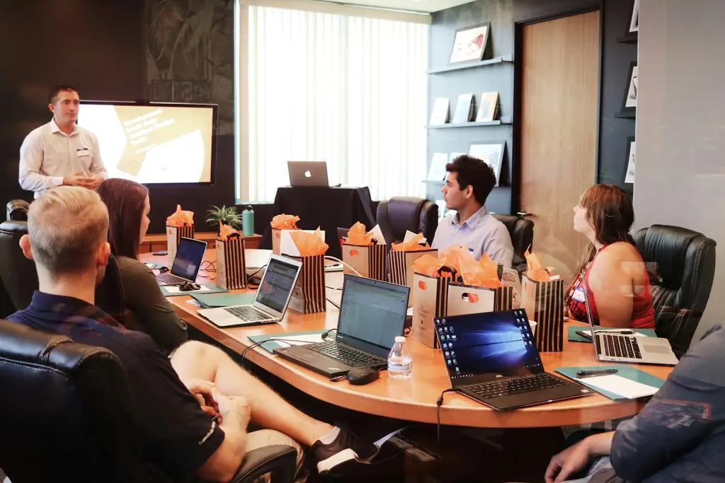 A presenter leading a discussion with colleagues seated around a meeting table with laptops